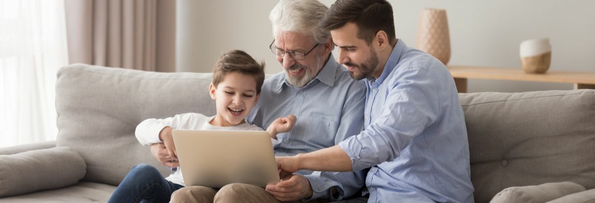 Dědeček, otec a syn společně sedí na gauči, drží laptop a smějí se.