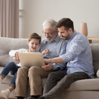 Dědeček, otec a syn společně sedí na gauči, drží laptop a smějí se.